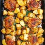 Close-up of baked chicken thighs and roasted potatoes garnished with herbs on a wooden table.