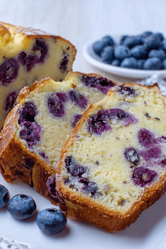 Sliced blueberry cream cheese loaf with blueberries and a cup of tea on a wooden cutting board.