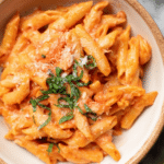 A colorful bowl of creamy tomato pasta garnished with parmesan cheese and basil on a wooden table.