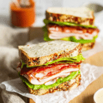 Delicious easy turkey club sandwich cut in half showing layers of turkey, bacon, avocado, and sun-dried tomatoes on a rustic wooden board with chips and salad.