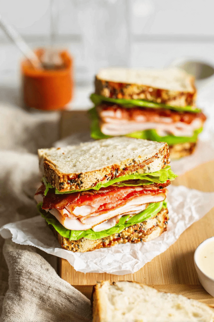 Delicious easy turkey club sandwich cut in half showing layers of turkey, bacon, avocado, and sun-dried tomatoes on a rustic wooden board with chips and salad.