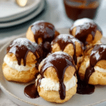 Freshly baked profiteroles drizzled with chocolate sauce on a plate.