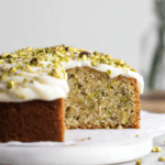 Slice of lemon pistachio cake with mascarpone frosting beside fresh lemons and pistachios.