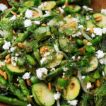 Vibrant sautéed spring vegetable salad with zucchini, asparagus, crumbled feta, and crushed pistachios on a rustic wooden table.