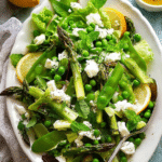 Colorful spring salad with asparagus, snow peas, baby cos lettuce, and goat cheese arranged on a wooden table.