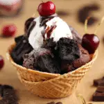 Close-up of a vegan chocolate brownie sundae in a waffle bowl topped with brownie chunks, coconut whip, chocolate shavings, and fresh cherries.