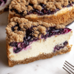 Slice of blueberry crumble cheesecake served on a plate with fresh blueberries, highlighting its creamy texture and crumble topping.