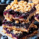 Freshly baked blueberry oatmeal bars displayed on a wooden board with blueberries scattered around.