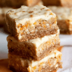 Delicious carrot cake bars topped with cream cheese frosting and cinnamon, served on a wooden table.