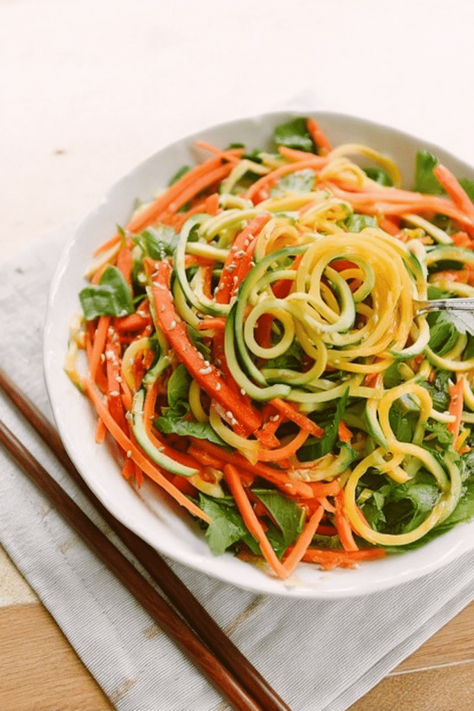 Cold spiralized sesame noodle salad with cucumber, carrot, zucchini, spinach, and almonds in a bowl, garnished with sesame seeds.