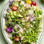 Everything Spring Green Salad with avocado, cherry tomatoes, and lemon basil vinaigrette on a wooden table.
