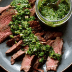 Garlic brown sugar flank steak topped with vibrant chimichurri sauce on a wooden board with fresh herbs and garlic.