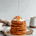 Stack of healthy carrot cake pancakes topped with almond butter, grated carrots, and maple syrup, served in a cozy breakfast setting.