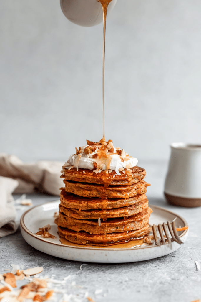 Stack of healthy carrot cake pancakes topped with almond butter, grated carrots, and maple syrup, served in a cozy breakfast setting.