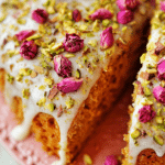 Persian Love Cake topped with rose petals and pistachios, served on decorative fabric with a cup of tea.