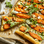 Roasted carrot puff pastry tart topped with arugula and walnuts, drizzled with honey, served on a rustic table.