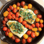 Plated roasted halibut with burst tomatoes, basil pesto, and lemon wedges.