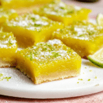 Vibrant lime bars dusted with powdered sugar and topped with lime zest on a wooden table.