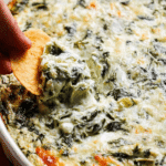 Baked spinach artichoke dip in a rustic dish garnished with herbs, served with crackers.