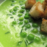 Bowl of fresh pea soup garnished with chives and croutons on a rustic wooden table.