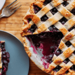 Freshly baked blueberry lattice pie on a wooden table with fresh blueberries and orange slices.