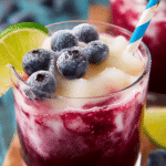 Refreshing boozy blueberry limeade slushie in a glass with lime wedges and blueberries on a vibrant summer background.