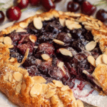 Cherry and almond galette topped with sliced almonds and ice cream on a wooden table, surrounded by fresh cherries.