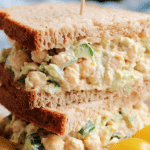 Chickpea salad sandwich stacked with fresh lettuce, tomato slices, and sprouts on a wooden table.