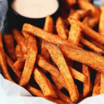 Crispy baked sweet potato fries in a bowl with a dipping sauce and herbs