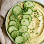 Bowl of dill pickle hummus with fresh vegetables and pita chips, garnished with dill and pickles