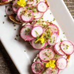 A plate of easy French buttered radish toasts topped with sliced radishes, chives, and edible flowers, ideal as an appetizer.