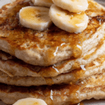 Fluffy banana pancakes stacked high with banana slices and maple syrup on a rustic table