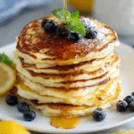 Fluffy lemon ricotta pancakes topped with fresh strawberries and syrup served on a white plate with lemon slices.