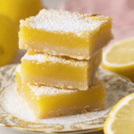 Delicious lemon bars dusted with powdered sugar on a white plate, garnished with lemon slices and mint leaves.