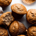 Delicious golden-brown banana muffins with melted butter and cinnamon on a wooden table, surrounded by fresh bananas and walnuts.