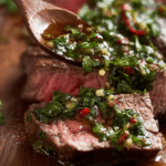 Grilled chimichurri steak with fresh herbs and lime slices on a wooden cutting board.