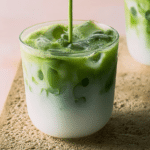 Iced matcha latte in a glass with ice, topped with matcha powder and surrounded by green tea leaves.