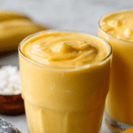 Mango coconut milk smoothie in a clear glass, garnished with mango slices and mint leaves on a wooden table.