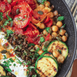 Mediterranean-style grain bowl with farro, chickpeas, lentils, zucchini, cherry tomatoes, avocado, and feta cheese on a wooden table.