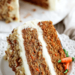 Slice of moist carrot cake with cream cheese frosting on a decorative plate, garnished with fresh carrots and walnuts.