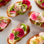 Radishes and butter crostini appetizer arranged on wooden board with chives and flake salt.