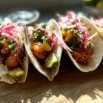Delicious saucy hot honey shrimp tacos topped with mango slaw, avocado, and pickled onions on a wooden board.