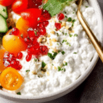 Savory cottage cheese bowl topped with cucumbers, bell peppers, tomatoes, and pistachios on a wooden table.