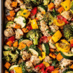 Sheet pan chicken with rainbow vegetables including broccoli, sweet potatoes, zucchini, and red bell peppers, topped with Parmesan cheese.