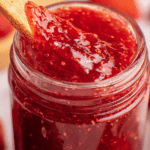 Jar of small batch strawberry jam surrounded by fresh strawberries and a wooden spoon in a rustic kitchen setting.