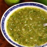 Bowl of spicy tomatillo salsa verde with fresh ingredients on a wooden table.