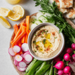 Colorful spring hummus platter with rainbow carrots, sugar snap peas, and sliced radishes, served with creamy hummus topped with olive oil and parsley.