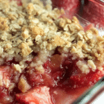 Close-up of homemade strawberry rhubarb crisp with golden crumble in a glass dish and fresh strawberries on the side.