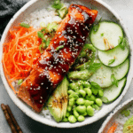 Vibrant teriyaki salmon bowl with salmon, brown rice, diced cucumbers, shredded carrots, shelled edamame, and avocado slices topped with green onions and sesame seeds.