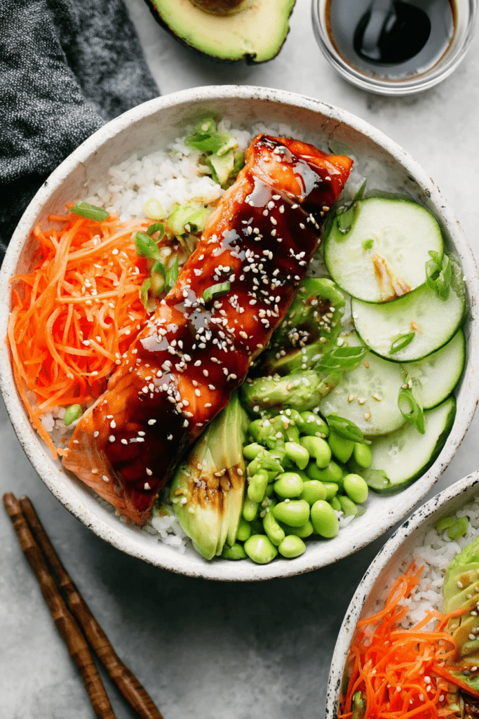 Vibrant teriyaki salmon bowl with salmon, brown rice, diced cucumbers, shredded carrots, shelled edamame, and avocado slices topped with green onions and sesame seeds.